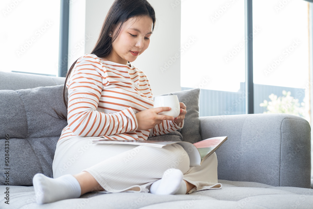 Young asian woman in casual clothes reading magazine, drinking tea on couch in modern interior Young asian woman in casual clothes reading magazine, drinking tea on couch in modern interior