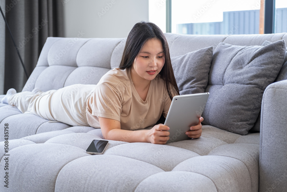Obraz premium beautiful young asian woman in casual cozy clothes using tablet sitting on couch at modern apartment