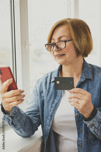 Happy senior woman holding bank credit card using smartphone, making internet payment, online shopping, booking hotel, order taxi via app, standing near window indoor. Online banking.Vertical. Mockup photo