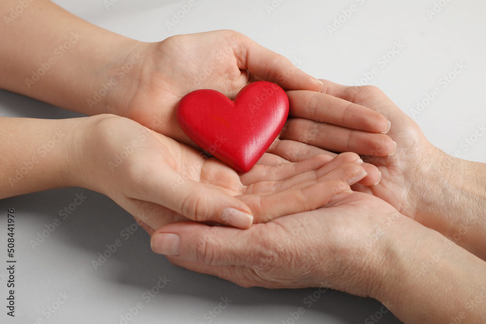 Young and elderly women holding red heart on light grey background, closeup