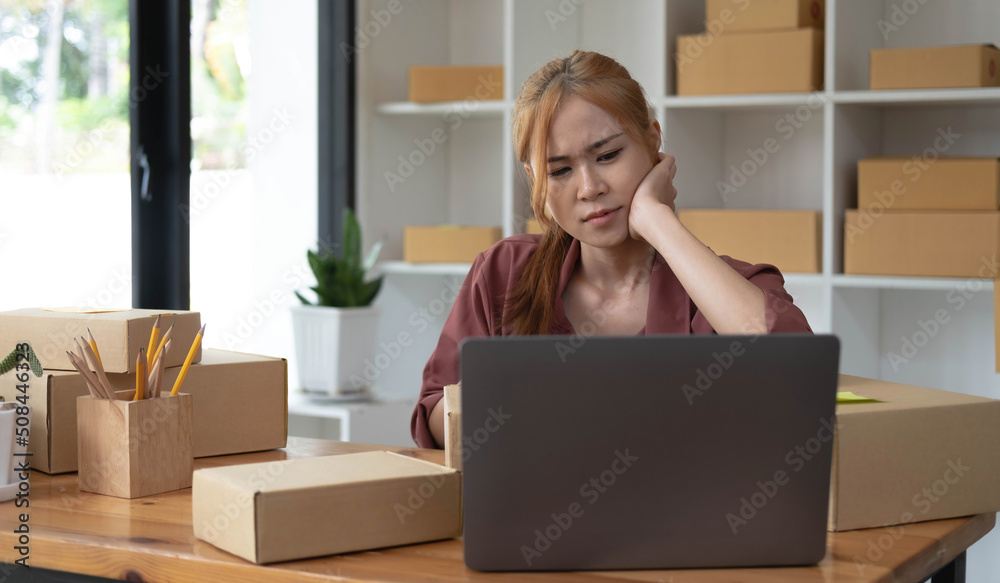 A portrait of young serious Asian woman working with laptop in the office full of packages and boxes stacking up, busy looking table, for SME, delivery, start up business and home office concept. A portrait of young serious Asian woman working with laptop in the office full of packages and boxes stacking up, busy looking table, for SME, delivery, start up business and home office concept.
