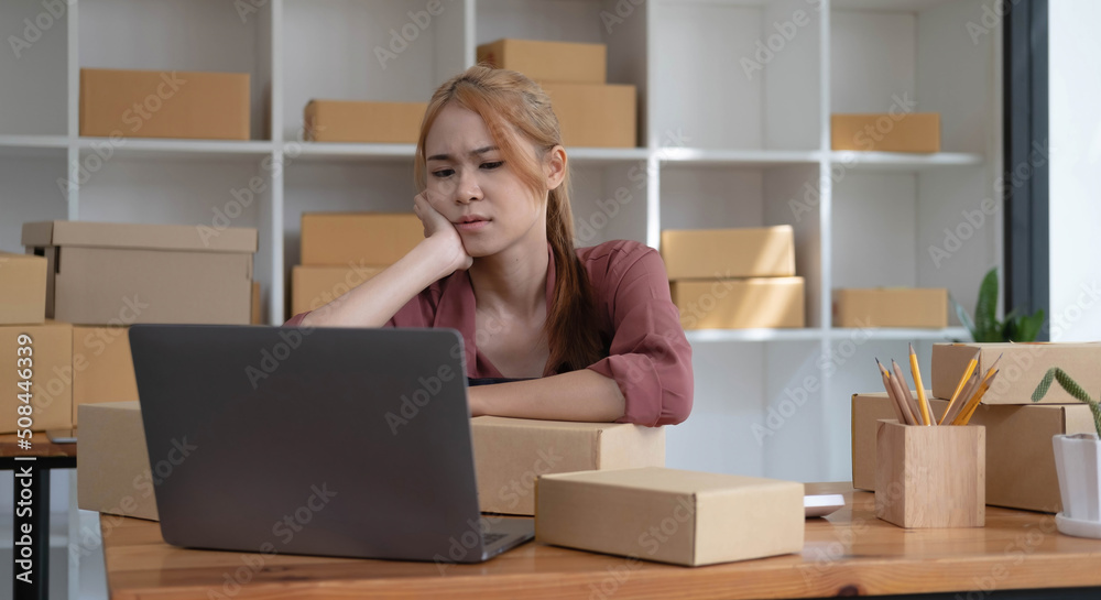 A portrait of young serious Asian woman working with laptop in the office full of packages and boxes stacking up, busy looking table, for SME, delivery, start up business and home office concept. A portrait of young serious Asian woman working with laptop in the office full of packages and boxes stacking up, busy looking table, for SME, delivery, start up business and home office concept.