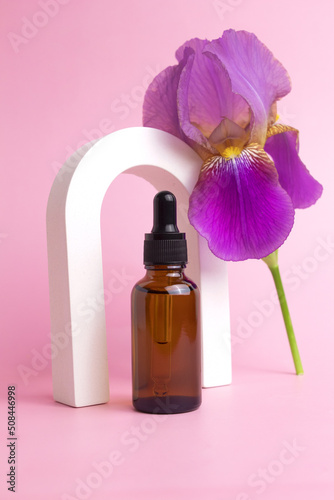 Serum with floral extracts for skincare. Face and body care spa concept. Nature cosmetics in glass bottle with a pipette and with iris flower on pink background
