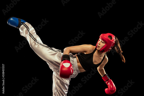 High kick. Attractive female boxer in boxing gloves and helmet training isolated on dark studio background. Sport, competition, hobby, results, success concept
