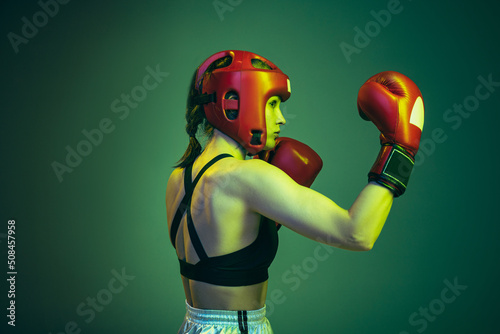 Portrait of professional female boxer in sports protective equipment posing isolated on green studio background in neon. Sport, competition, hobby, results, success concept