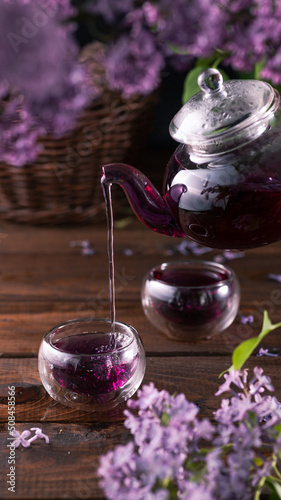 Purple tea is poured from a glass teapot into cups. Lilac flowers are all around
