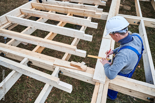 Man worker building wooden frame house on pile foundation. Carpenter using angle for measuring wooden planks and making marks with pencil. Carpentry concept.