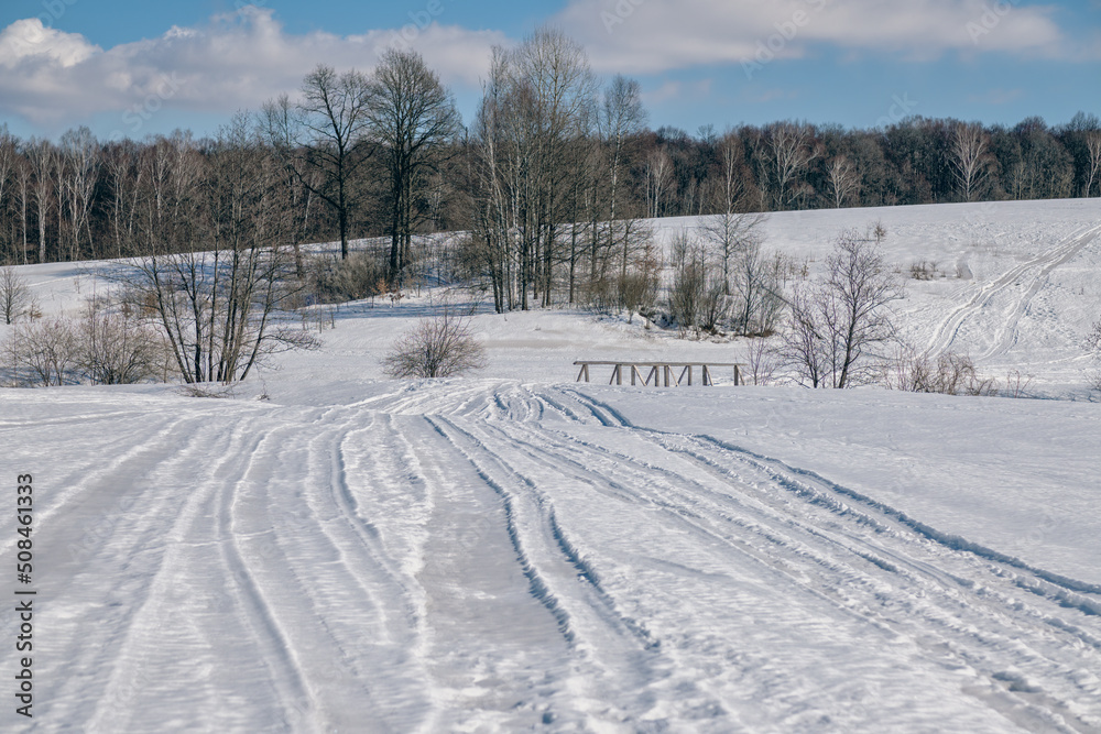 Wooden bridge between the slopes of the hilly area in winter with tracks on the snow and trees in the background.