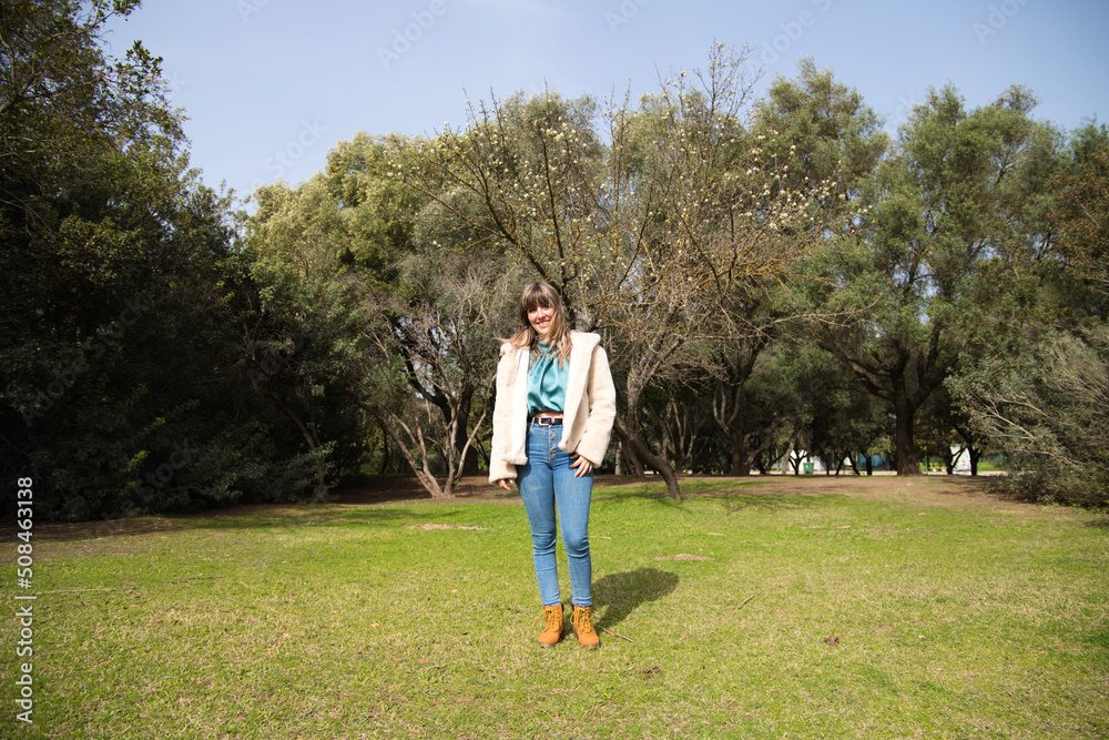 young and beautiful blonde woman dressed in jeans, coat with white cap and boots is walking in the park and is having her photo taken while enjoying the sunny day among the trees. Holiday concept. young and beautiful blonde woman dressed in jeans, coat with white cap and boots is walking in the park and is having her photo taken while enjoying the sunny day among the trees. Holiday concept.