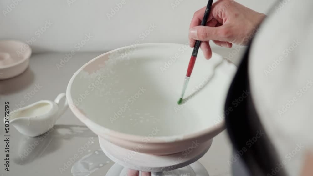 Female hands paint a ceramic bowl with a brush. Women's hands apply white paint on a blue base, the artist works in a pottery studio with a potter's wheel. Close up. Female hands paint a ceramic bowl with a brush. Women's hands apply white paint on a blue base, the artist works in a pottery studio with a potter's wheel. Close up.