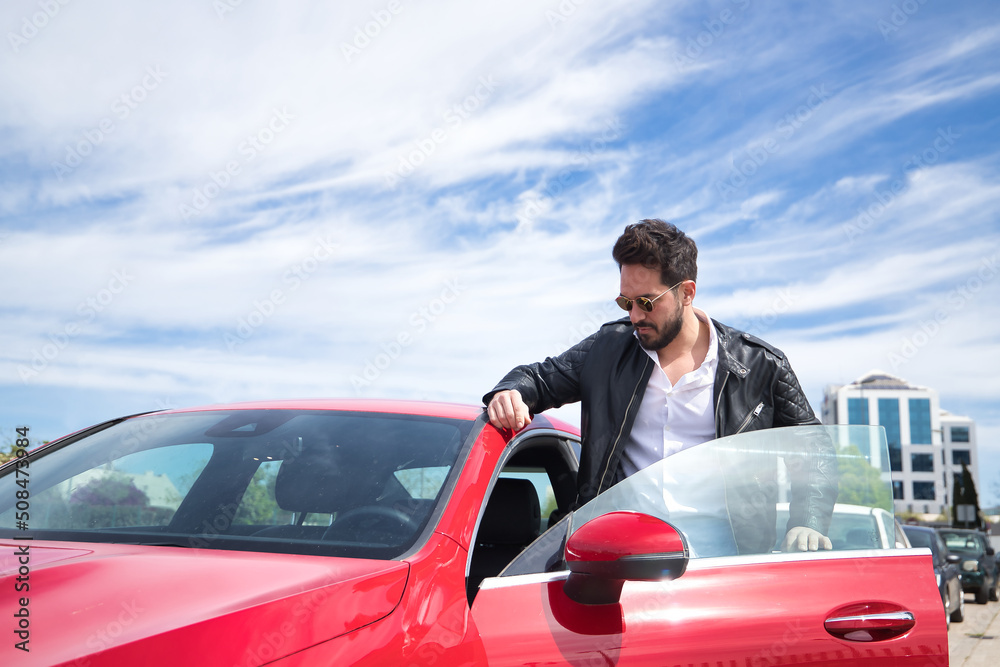 Handsome young man with beard, sunglasses, leather jacket and white shirt, getting into his red sports car. Concept beauty, fashion, trend, luxury, motor, sports. Handsome young man with beard, sunglasses, leather jacket and white shirt, getting into his red sports car. Concept beauty, fashion, trend, luxury, motor, sports.