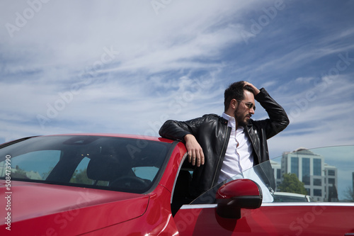 Handsome young man with beard, sunglasses, leather jacket and white shirt, leaning on the roof of his red sports car, while touching his hair. Concept beauty, trend, luxury, motor, sports, hairdresser
