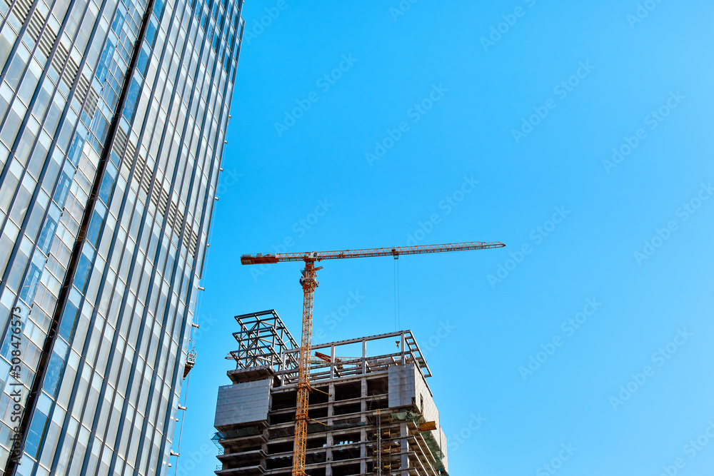 Fototapeta premium The base of a building under construction and a yellow tower crane along it next to part of a glass skyscraper against a clear blue sky in the light of the sun