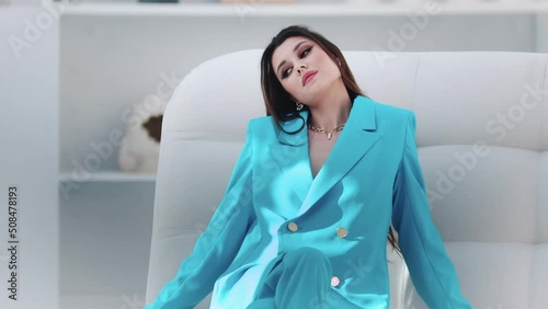 Young fashionable woman in blue blazer sits on the couch in white studio and posing for the camera photo