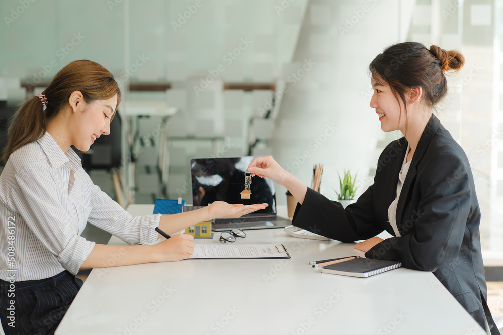 Fototapeta premium A saleswoman checks the lease and holds a small gray model of house and house keys in preparation for meeting customers to sign the lease. Real estate leasing concept.