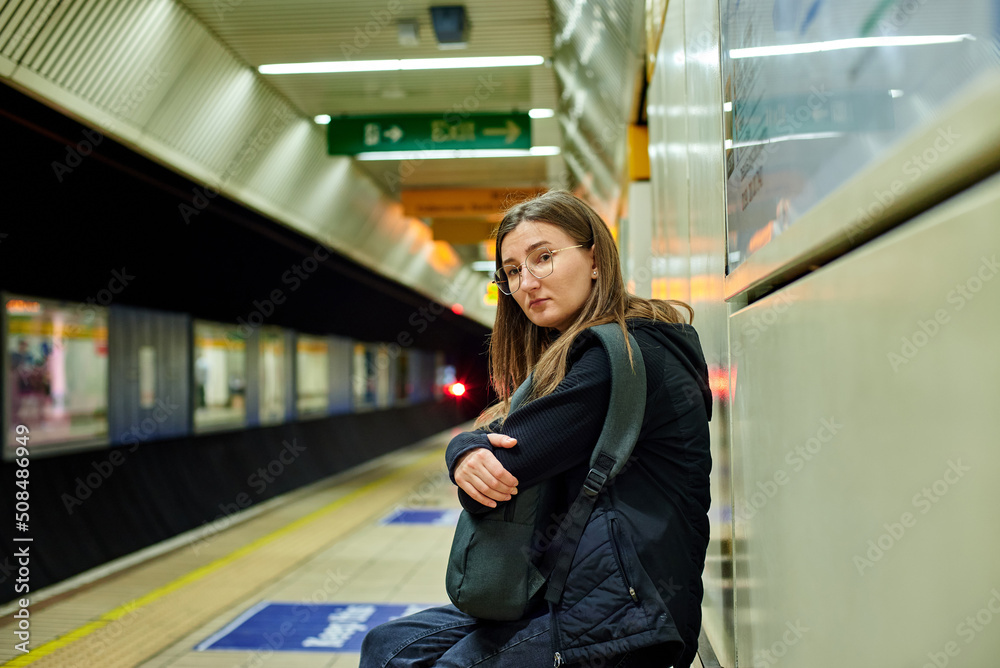 A girl with glasses is waiting for a train in the subway, sitting on a bench and hugging her backpack. A girl with glasses is waiting for a train in the subway, sitting on a bench and hugging her backpack.