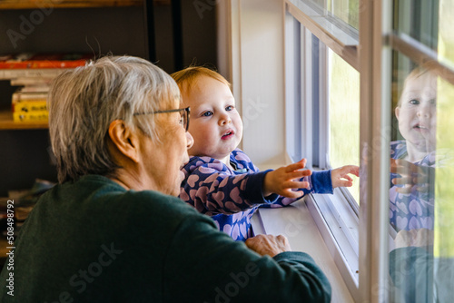 Asian grandmother looks out of a window with her mixed race granddaughter pointing 