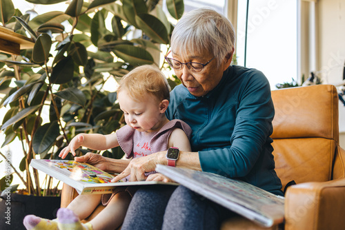 Senior asian woman reads a book with her mixed-race granddaughter at home in front of a plant