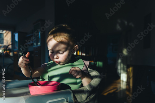 A baby wearing a bib eats out of a bowl in dramatic lighting