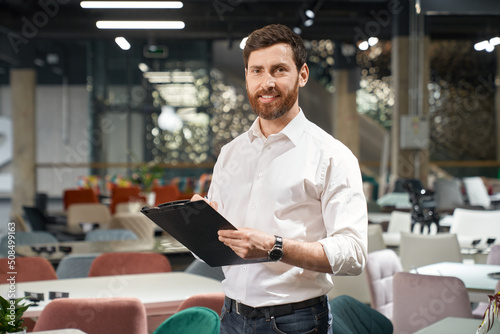 Front view of brunette male with beard standing, holding folder in furniture store. Designer, seller, architect writing, designing, looking at camera. Concept of furniture. photo