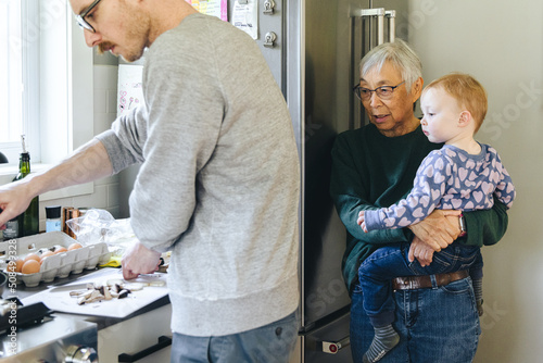 Asian grandmother holds her granddaughter while her son-in-law cooks dinner in the kitchen