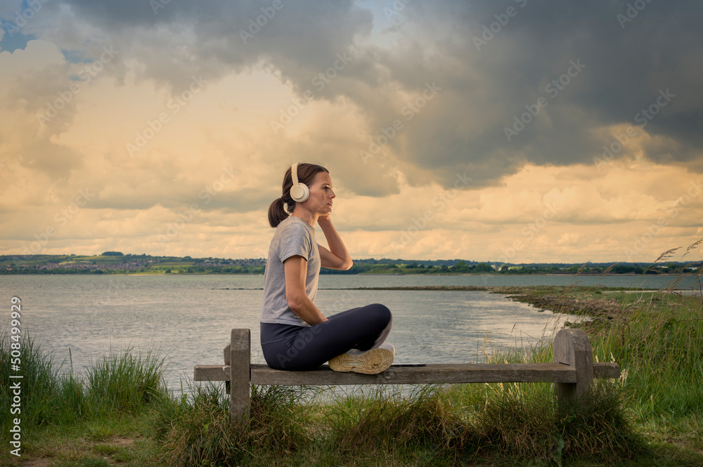 Single woman sitting on a bench beside water listening to music on her headphones.