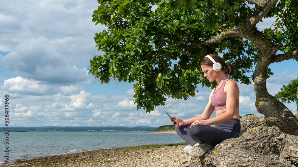 woman sitting under a tree by water listening to podcast or music in the sun.