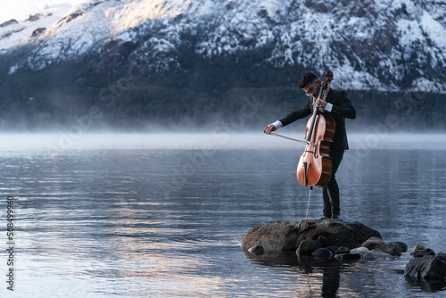 Musician playing cello by the lake with snow capped mountains on the back