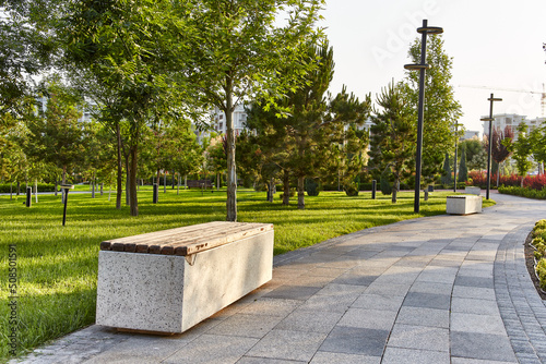 White stone and wood bench on the sidewalk in the park with fresh green grass and trees around at dawn light with houses in the background