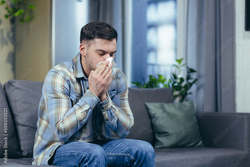 Feeling unwell. A young handsome man is sitting on the couch at home, is sick, feels cold, wipes his nose with a napkin, has a runny nose. Feeling unwell. A young handsome man is sitting on the couch at home, is sick, feels cold, wipes his nose with a napkin, has a runny nose.