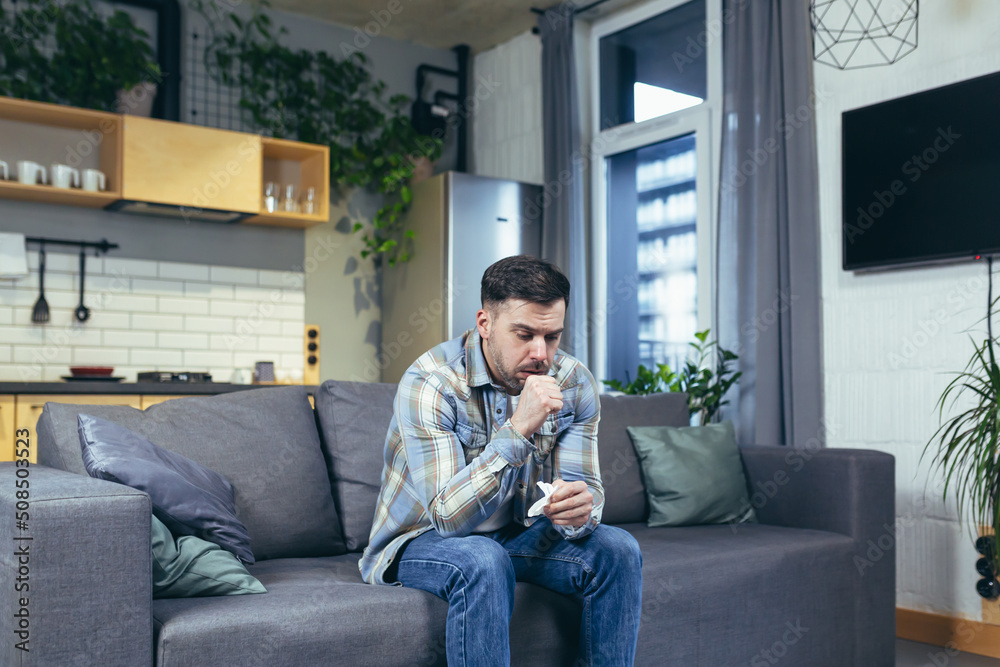 Feeling unwell. A young handsome man is sitting on the couch at home, is sick, feels cold, wipes his nose with a napkin, has a runny nose.