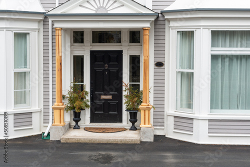 A black metal formal looking door with two flower pots on both sides. The entrance to the grey house has glass windows and wood columns. The siding is a wood clapboard with white trim.  © Dolores  Harvey