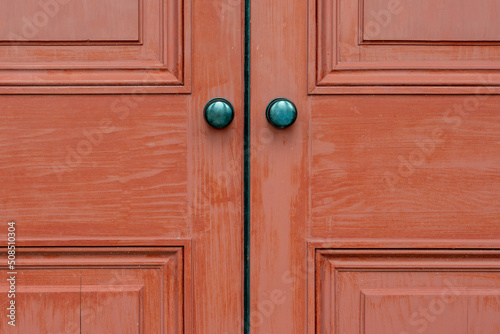 Two red wooden doors to a house with two vintage round metal doorknobs. The exterior panel doors show wood grain and worn patterns around the handles. The retro style of doors is hardwood. © Dolores  Harvey