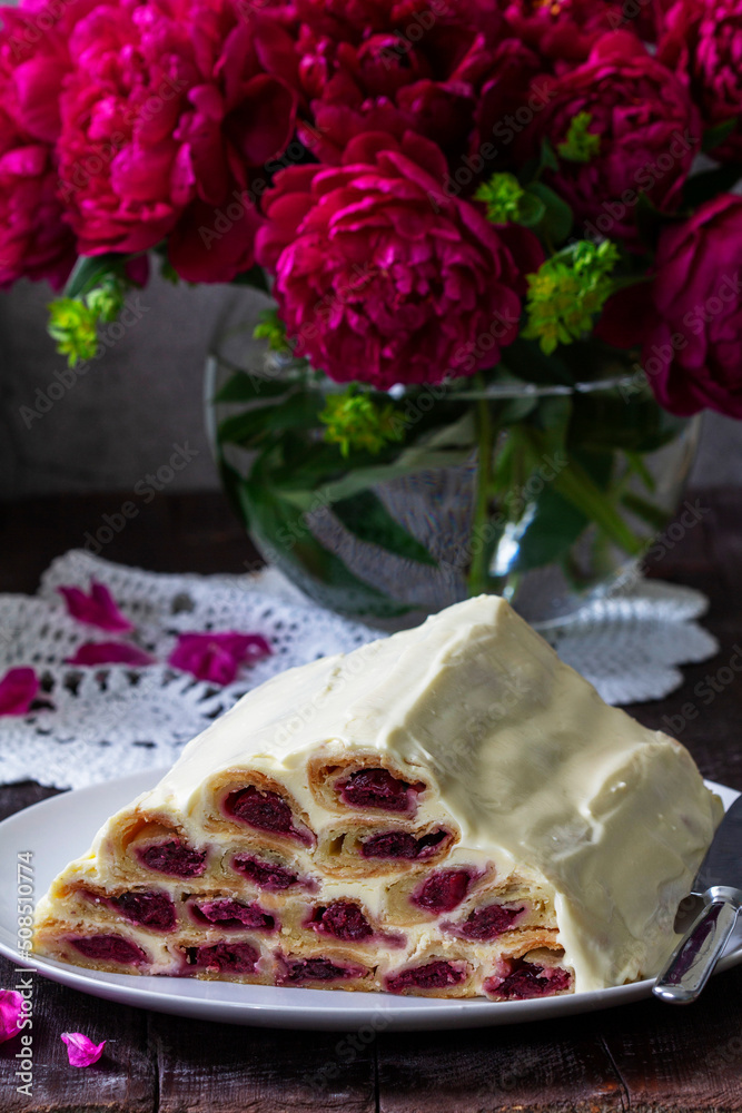 Cake Monastic hut with cherry filling and sour cream, a bouquet of peonies on a wooden table.