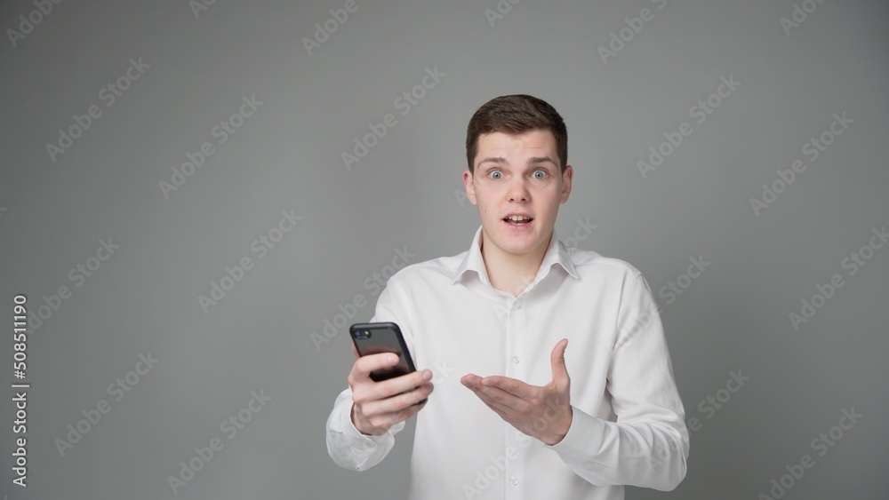 A surprised young guy looks at the screen of his smartphone. On a gray background. A surprised young guy looks at the screen of his smartphone. On a gray background.