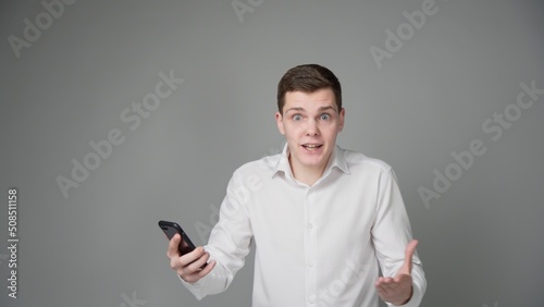 A surprised young guy looks at the screen of his smartphone. On a gray background. © Vlamin