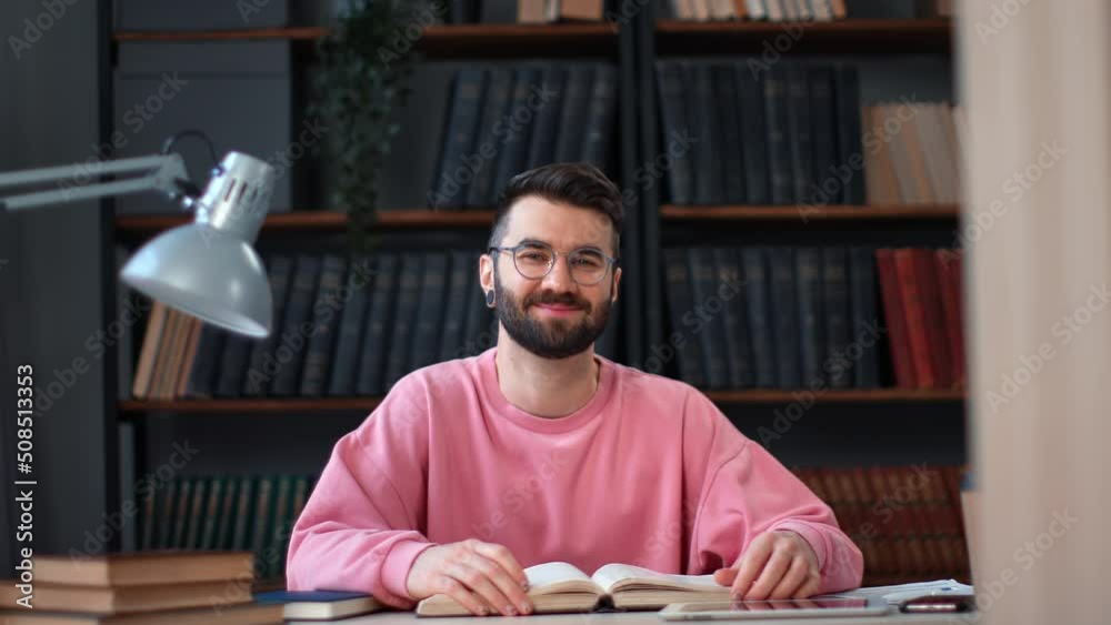 Portrait smiling male student in pink sweatshirt posing at library desk open book enjoy education Portrait smiling male student in pink sweatshirt posing at library desk open book enjoy education