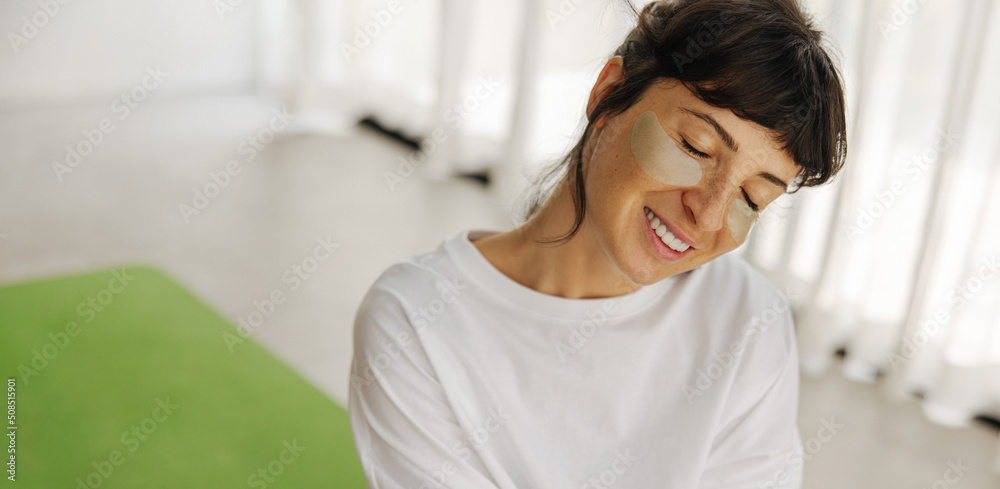 Close-up of cute young caucasian woman enjoying patches smiling with her eyes closed indoors. Brunette in white t-shirt is doing cosmetic procedures. Health care lifestyle concept