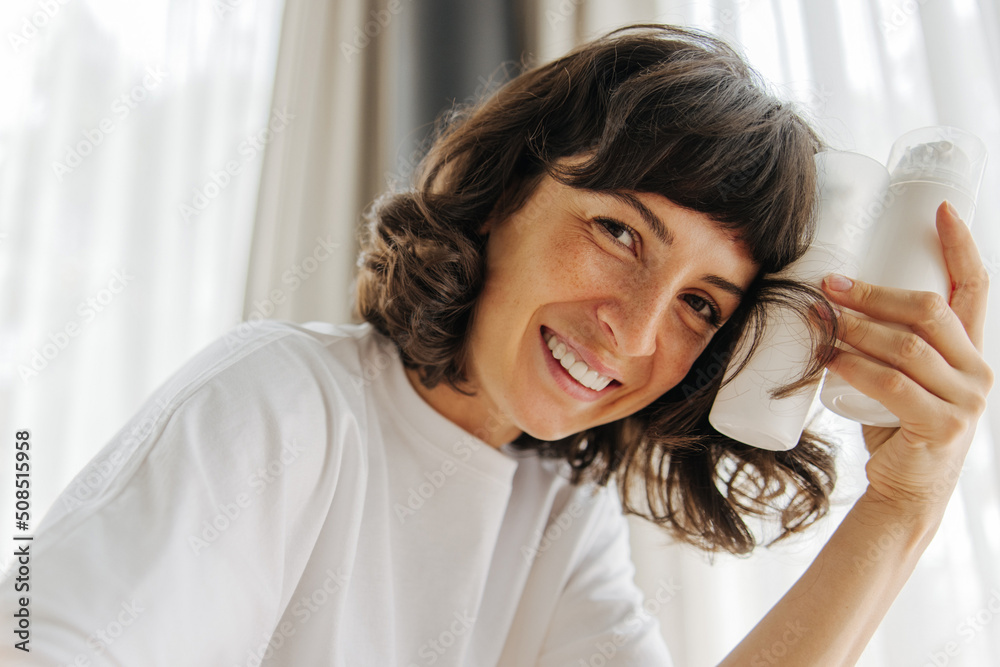 Fototapeta premium Influential young caucasian girl is looking at camera holding jars of facial foam indoors. Brunette wears white t-shirt in morning at home. Female beauty rituals, skin care cosmetic products concept