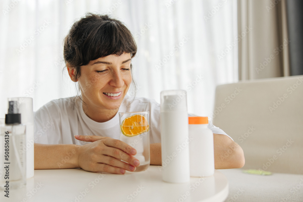 Beautiful young caucasian girl sits at table with care cosmetics, drinks water with lemon. Brunette wears white t-shirt and chooses cosmetic product. Health concept Beautiful young caucasian girl sits at table with care cosmetics, drinks water with lemon. Brunette wears white t-shirt and chooses cosmetic product. Health concept