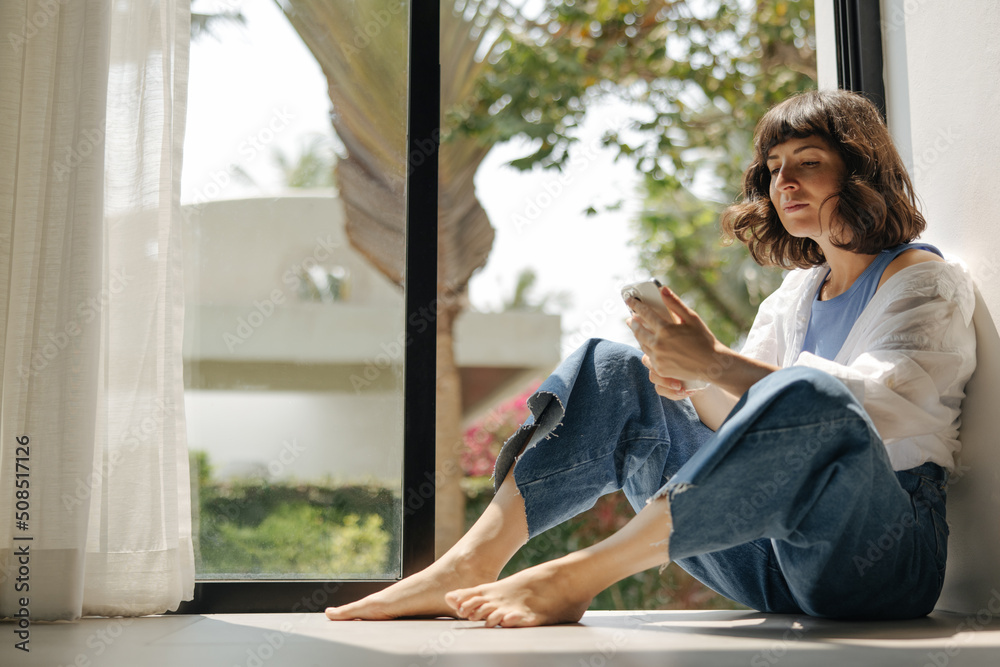 Cute young caucasian woman is using modern smartphone sitting by window in morning. Brunette with short haircut wears shirt and jeans. Daylight saving time concept Cute young caucasian woman is using modern smartphone sitting by window in morning. Brunette with short haircut wears shirt and jeans. Daylight saving time concept