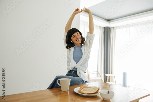 Happy young caucasian brunette woman closing her eyes sits in interior of kitchen at breakfast. Girl in casual clothes eats delicious pancakes, keeping lifestyle. Good morning concept