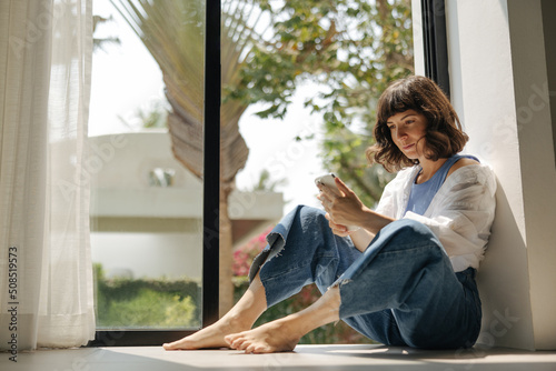 Pleasant young caucasian woman plays game on phone sitting by window in warm weather. Brunette wears casual clothes at home. Technology concept Free Download Pleasant young caucasian woman plays game on phone sitting by window in warm weather. Brunette wears casual clothes at home. Technology concept - GFXLoad