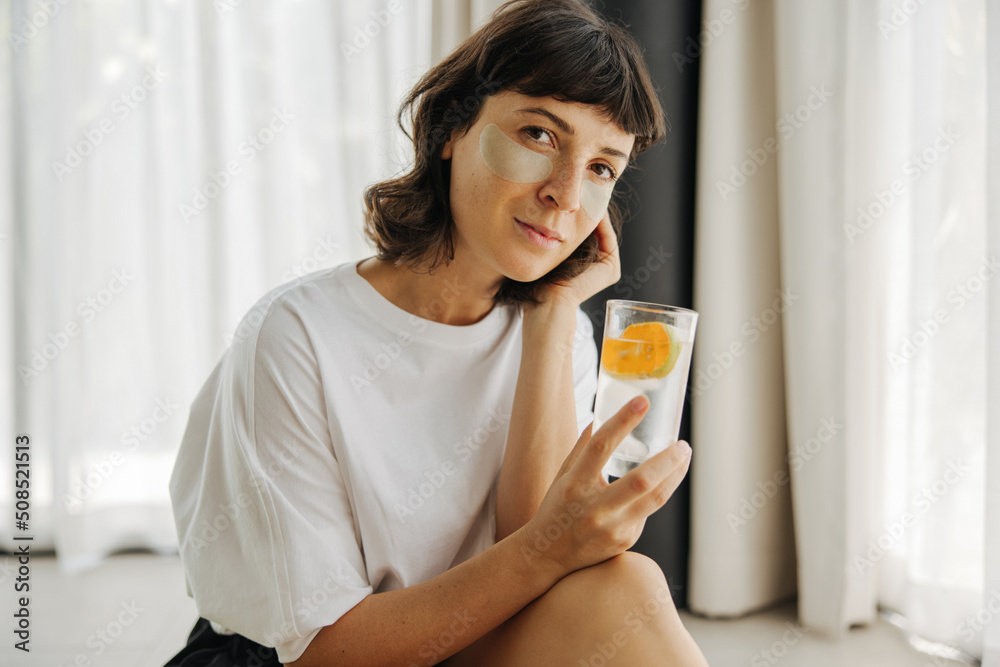 Beautiful young caucasian girl with patches on face and looking at camera sitting indoors. Brunette in white t-shirt holds glass of clean water with citrus. Skin care and health concept Beautiful young caucasian girl with patches on face and looking at camera sitting indoors. Brunette in white t-shirt holds glass of clean water with citrus. Skin care and health concept