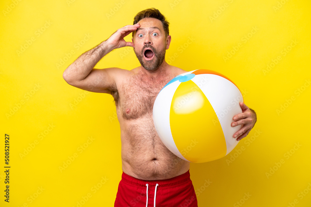 Middle age caucasian man holding beach ball isolated on yellow background with surprise expression