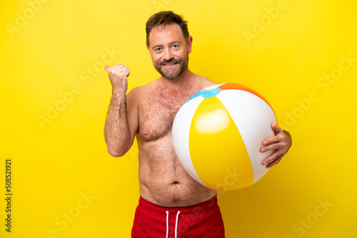 Middle age caucasian man holding beach ball isolated on yellow background pointing to the side to present a product