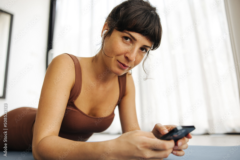 Calm young caucasian woman is lying on mat browsing smartphone at home. Girl in brown top and leggings looks at camera. Concept of rest, relaxation, technology and lifestyle. Calm young caucasian woman is lying on mat browsing smartphone at home. Girl in brown top and leggings looks at camera. Concept of rest, relaxation, technology and lifestyle.