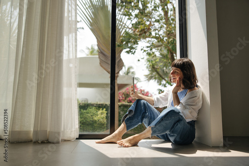 Friendly young caucasian woman waving at screen of smartphone communicating via video communication sitting by window. Brunette wears casual clothes at home. Lifestyle, different emotions, concept.