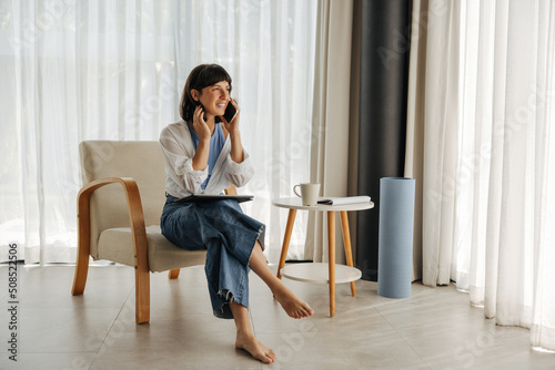 Smiling young caucasian woman talking on phone sitting on easy chair indoors. Brunette wears casual clothes at home. Cell phone usage concept © Look!