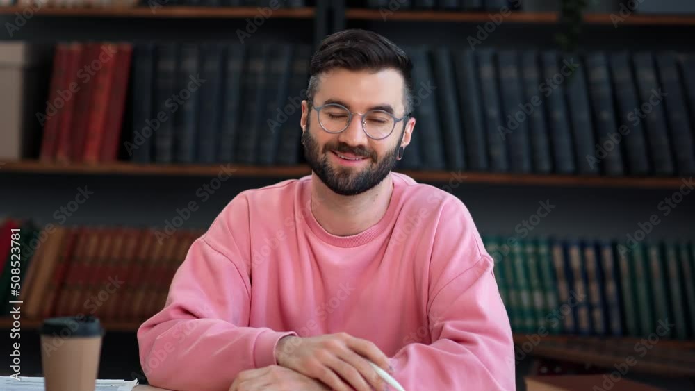 Smiling casual male blogger in pink sweatshirt speaking webinar channel auditorium at library desk